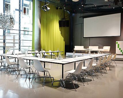 In Space04 at Kunsthaus Graz, white tables with armchairs stand in front of the podium in the background. The screen for projections is lowered. On the podium are white couches and a lectern.