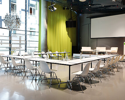 Im Space04 im Kunsthaus Graz stehen weiße Tische mit Sesseln vor dem Podium im Hintergrund. Die Leinwand für Projektionen ist heruntergelassen. Auf dem Podium stehen weiße Couchen sowie ein Rednerpult.