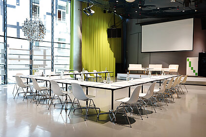Im Space04 im Kunsthaus Graz stehen weiße Tische mit Sesseln vor dem Podium im Hintergrund. Die Leinwand für Projektionen ist heruntergelassen. Auf dem Podium stehen weiße Couchen sowie ein Rednerpult.