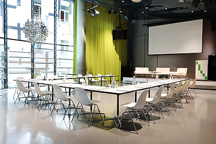 In Space04 at Kunsthaus Graz, white tables with armchairs stand in front of the podium in the background. The screen for projections is lowered. On the podium are white couches and a lectern.