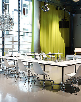 In Space04 at Kunsthaus Graz, white tables with armchairs stand in front of the podium in the background. The screen for projections is lowered. On the podium are white couches and a lectern.