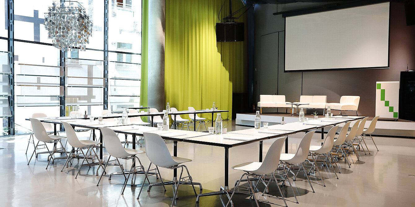 Im Space04 im Kunsthaus Graz stehen weiße Tische mit Sesseln vor dem Podium im Hintergrund. Die Leinwand für Projektionen ist heruntergelassen. Auf dem Podium stehen weiße Couchen sowie ein Rednerpult.