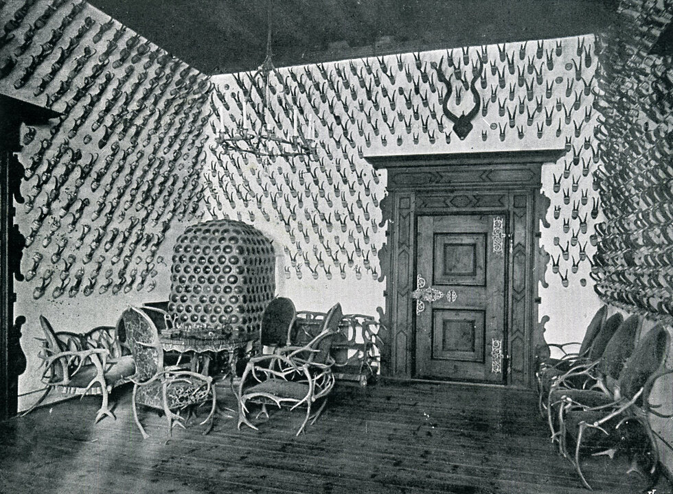 Alte Fotografie von einem Zimmer. An den Wänden hängen von oben bis unten Geweihe. In der linken Ecke steht ein Ofen davor sind Möbelstücke aus Geweihen zusehen.
