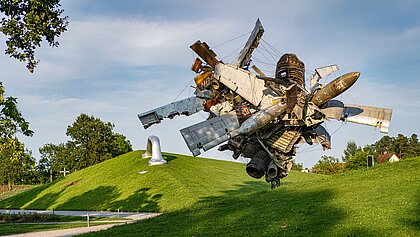 Blick vom Wegesrand auf die Skulptur "Airplane Parts & Hills" von Nancy Rubins und "Die Erdkugel als Koffer" von Peter Weibel in der Ferne