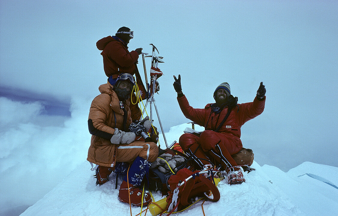 Alte Fotografie von drei Männern auf einem verschneiten Gipfel. Vor ihnen liegt ihre Ausrüstung. Ein Mann streckt die Hände in die Höhe und zeigt das "Peace"-Zeichen. Im Hintergrund sind ausschließlich Wolken und Nebel zusehen.  