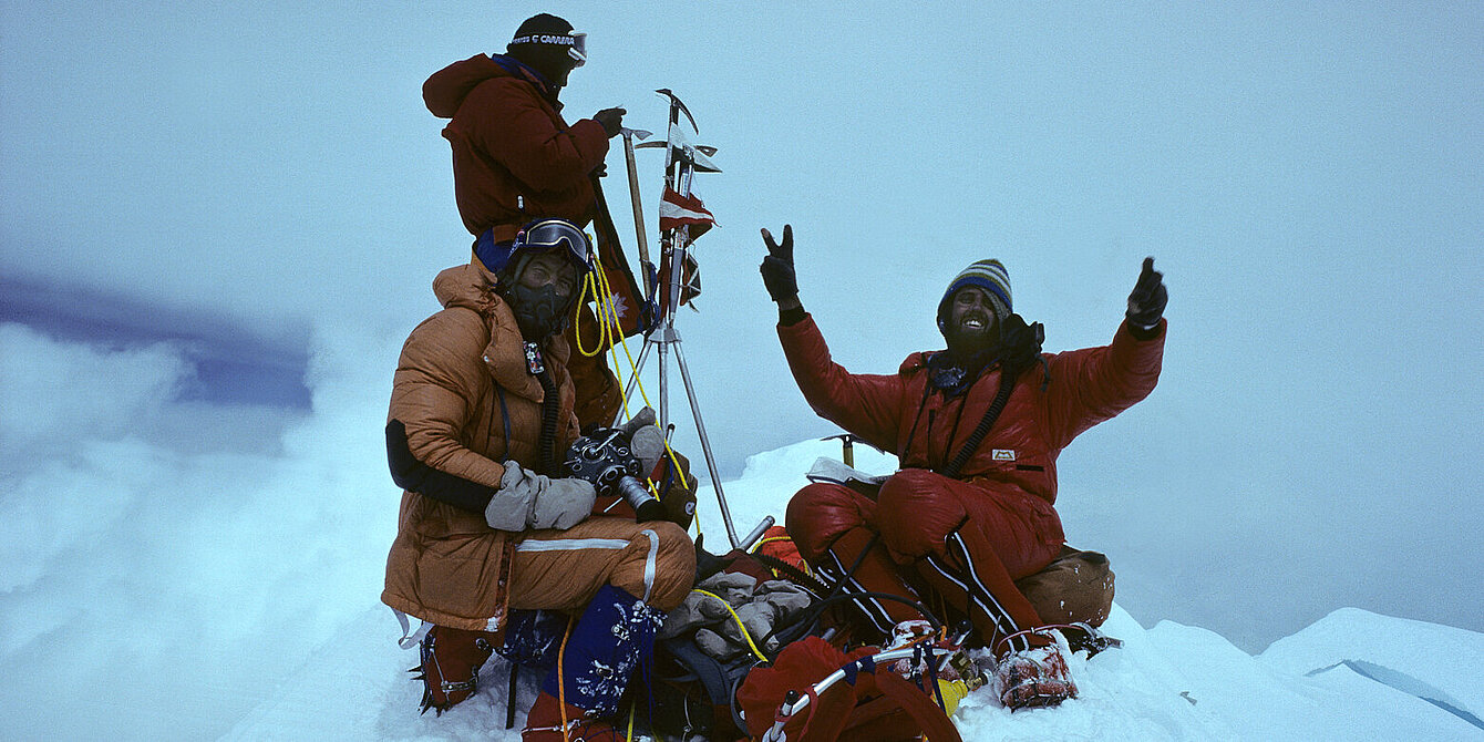 Alte Fotografie von drei Männern auf einem verschneiten Gipfel. Vor ihnen liegt ihre Ausrüstung. Ein Mann streckt die Hände in die Höhe und zeigt das "Peace"-Zeichen. Im Hintergrund sind ausschließlich Wolken und Nebel zusehen.  