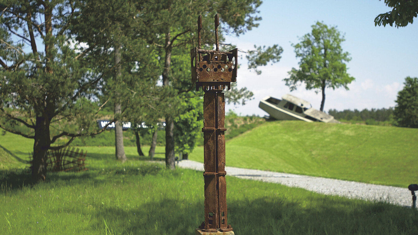 Gerhard Moswitzers Metallskulptur steht auf einem Rasenstück des Berggartens. Im Hintergrund sieht man verschwommen weitere Skulpturen. 