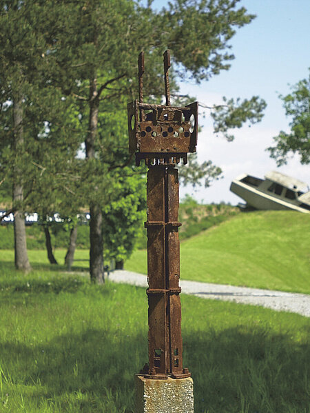 Gerhard Moswitzers Metallskulptur steht auf einem Rasenstück des Berggartens. Im Hintergrund sieht man verschwommen weitere Skulpturen. 