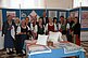 A group of elderly women in traditional cloths stands in a room. In front of and behind them knitted and embroidered goods are presented.