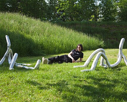Die Künstlerin Barbara Kapusta liegt zwischen ihren Skulpturen "Second and Four" im sommerlich grünen Skulpturenpark. 