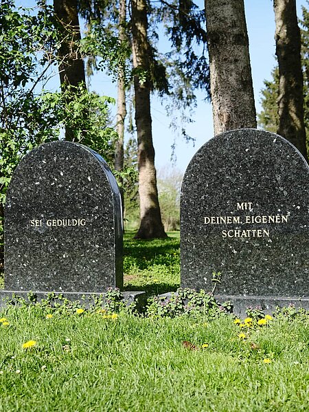 Zwei Grabsteine aus glattem Stein. Die Inschrift des ersten: Sei Geduldig". Die Inschrift des Zweiten: "Mit deinem eigenen Schatten".