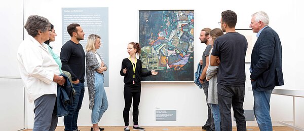 Eine Mitarbeiterin der Neuen Galerie spricht vor einer Gruppe von Besucher:innen, die vor einem Gemälde von Egon Schiele mit dem Titel "Stadtende/Häuserbogen III" steht.