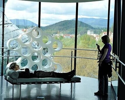 Ein Mann liegt auf einer Recamiere in der Needle im Kunsthaus Graz. Eine Frau macht ein Foto davon.