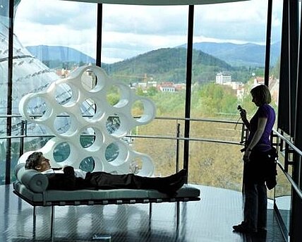 A man lies on a recamiere in the Needle in the Kunsthaus Graz. A woman takes a photo of it.