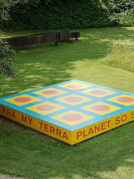 Die Skulptur ist eine rechteckige Plattform mit der Aufschrift "TERRA MY TERRA/PLANET SO SWEET/I CAN FEEL YOU/UNDER MY FEET". Die Plattform ist bemalt mit geometrischen Formen in blau, gelb und rot.