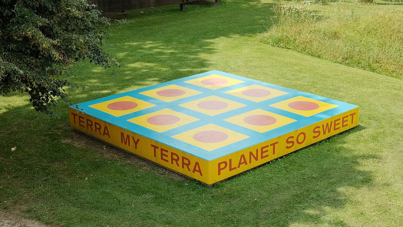 Die Skulptur ist eine rechteckige Plattform mit der Aufschrift "TERRA MY TERRA/PLANET SO SWEET/I CAN FEEL YOU/UNDER MY FEET". Die Plattform ist bemalt mit geometrischen Formen in blau, gelb und rot.