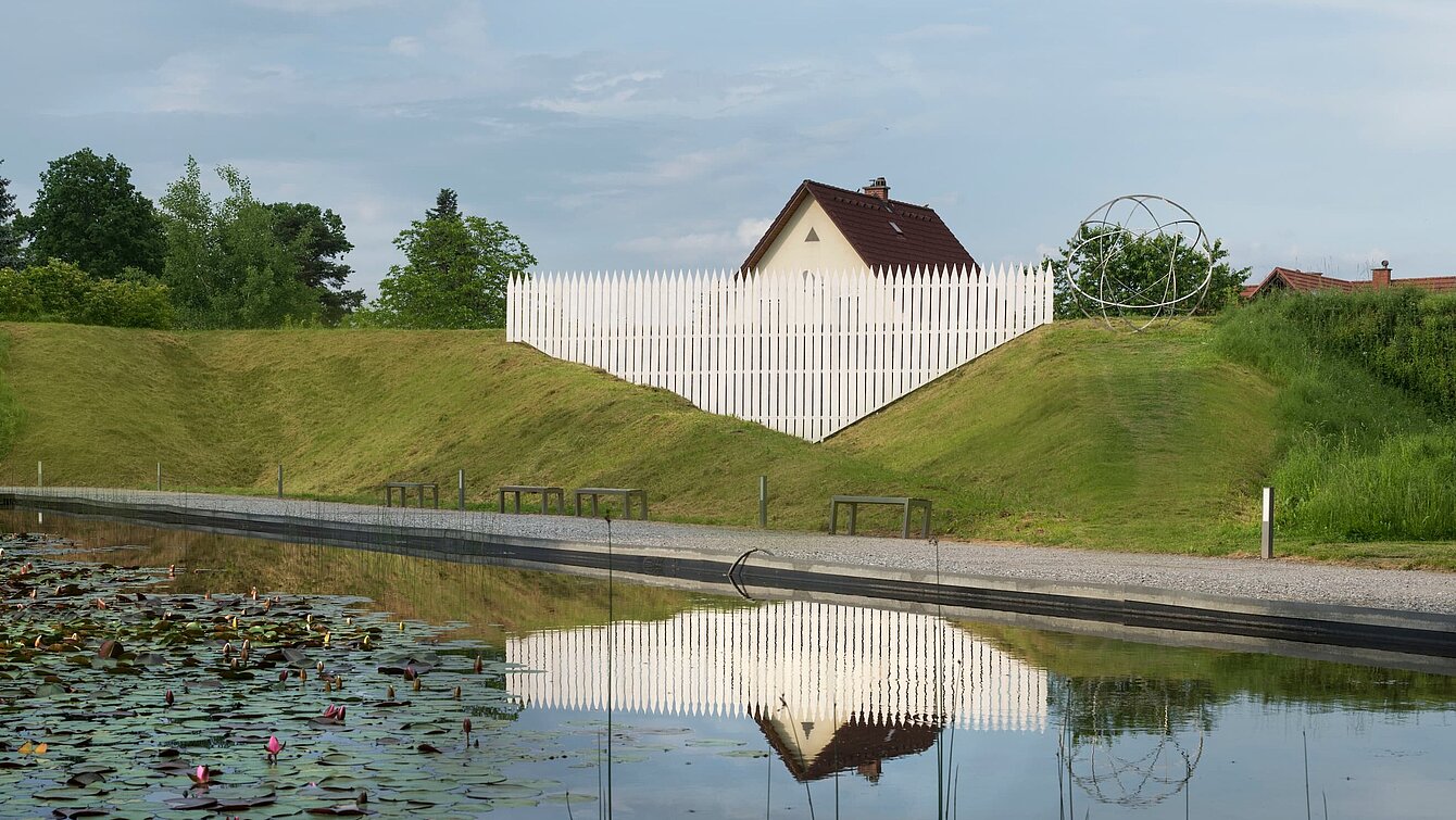 An oversized garden fence that incorporates the surroundings outside the sculpture park. The roof of a typical detached house protrudes from behind the wall of the park, appearing strangely small, almost sunken, due to the oversized fence.