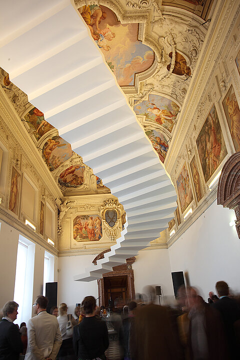 Zu sehen ist der Marmorsaal in dem eine Gruppe von Menschen steht. An der Decke sind die Fresken zu sehen, darunter hängt in der Luft schwebend eine weiße Treppe.