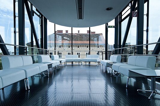 Blick in Richtung Süden in der Needle im Kunsthaus Graz. Links und rechts stehen weiße Sitzcouchen, im Hintergrund sind man das Eckgebäude Lendkai/Südtiroler Platz.