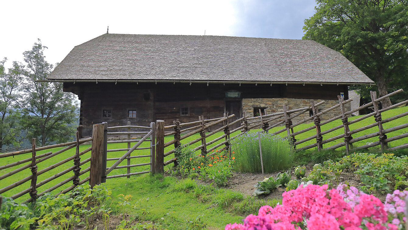 Blick vom Garten mit blühenden Blumen über den Bänderzaun zum Geburtshaus.