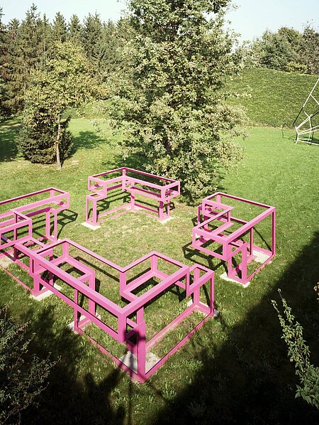 The sculpture is a large walk-in structure made of pink painted metal. The ground plan is square, with an "entrance" on each side.