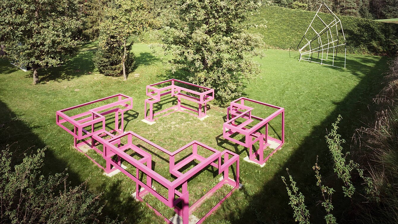 The sculpture is a large walk-in structure made of pink painted metal. The ground plan is square, with an "entrance" on each side.