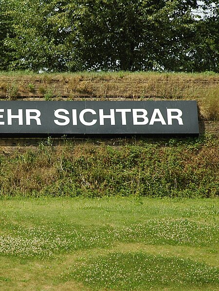 White lettering on a black background inside the wall of the sculpture park with the inscription "Nicht mehr sichtbar" [no longer visible]. On the other side of the wall on the grounds of the Schwarzl leisure center is the counterpart with the inscription "Noch nicht sichtbar" [not yet visible].