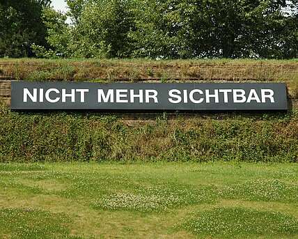 Weißer Schriftzug auf schwarzem Untergrund innen am Wall des Skulpturenparks mit der Aufschrift "nicht mehr sichtbar". Auf der anderen Seite des Walls auf dem Gelände des Schwarzl Freizeitzentrums befindet sich das Gegenstück mit der Aufschrift "noch nicht sichtbar".