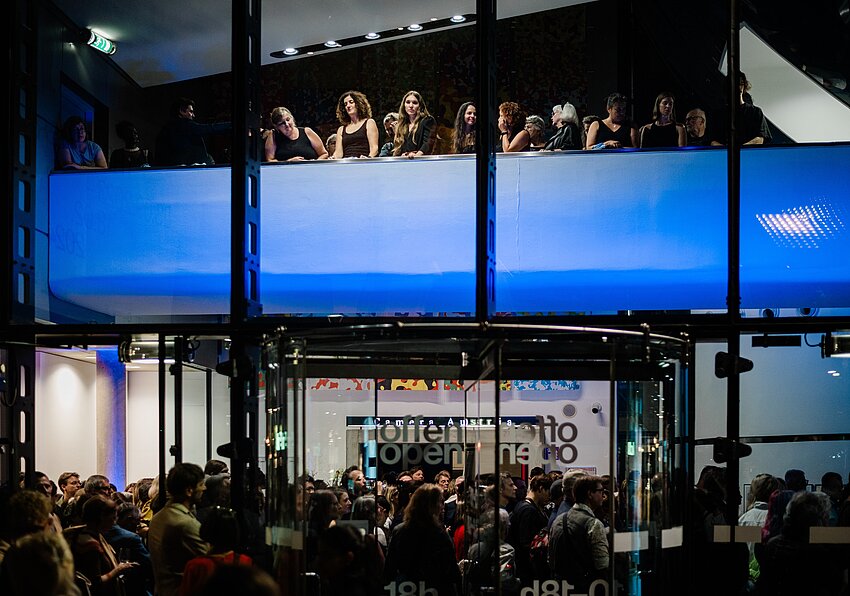 Das Kunsthaus Graz wird nachts von außen fotografiert. Durch die Glasfassade sieht man das blau beleuchtete Foyer des Kunsthauses. Eine große Menschenmenge versammelt sich im Foyer und ein Chor steht über der Menschenmenge auf einer balkonartigen Brücke. 
