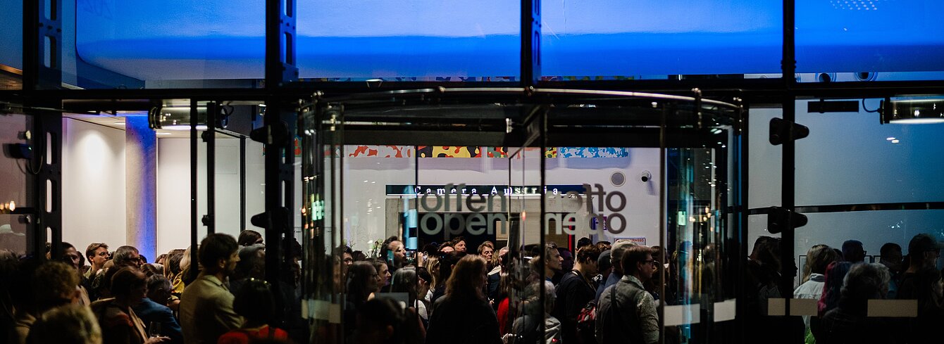 Das Kunsthaus Graz wird nachts von außen fotografiert. Durch die Glasfassade sieht man das blau beleuchtete Foyer des Kunsthauses. Eine große Menschenmenge versammelt sich im Foyer und ein Chor steht über der Menschenmenge auf einer balkonartigen Brücke. 