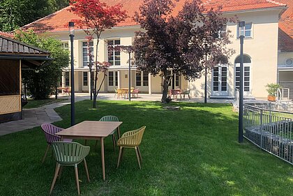 Blick Richtung Heimatsaal im Duftgarten. Im Vordergrund ist ein Tisch mit Sesseln, seitlich sieht man den Pavillon.