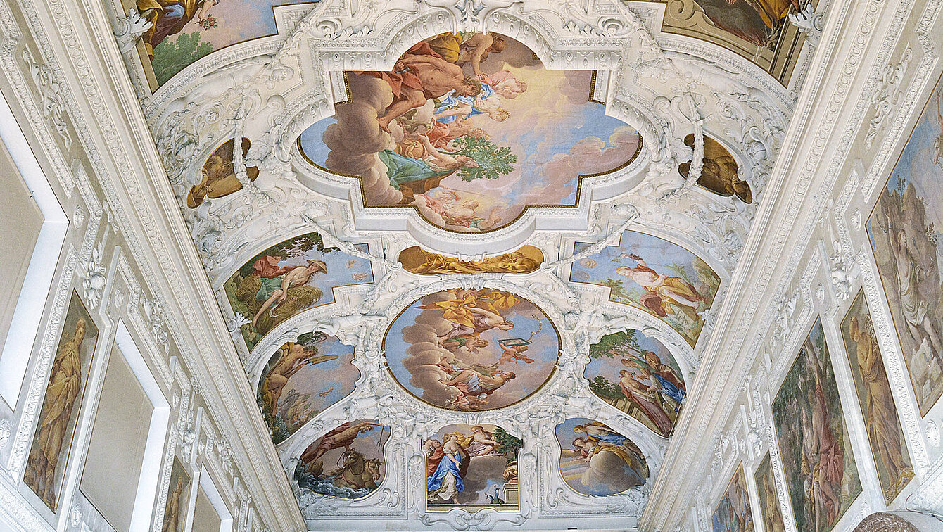 Die Decke einer großen Halle, die mit weißen Stuck und bunten Malereien verziert ist. Die Malereien zeigen Figuren aus Sagen und Religionen. 