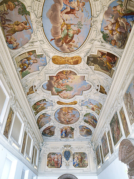 Die Decke einer großen Halle, die mit weißen Stuck und bunten Malereien verziert ist. Die Malereien zeigen Figuren aus Sagen und Religionen. 