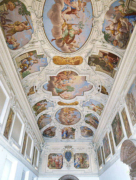 The ceiling of a large hall decorated with white stucco and colorful paintings. The paintings depict figures from legends and religions.