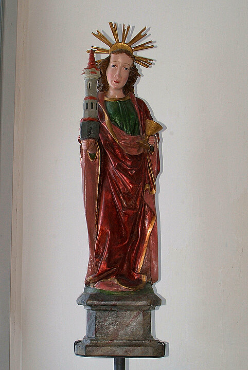 Wooden statue of a woman with a red robe and a golden halo. In her hand she holds a small gray tower with a red pointed roof.