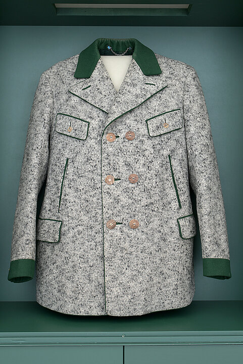 Photo of a coat hanging in a display case. The coat is made of gray loden and has a green collar. There are three rows of double buttons at the front.
