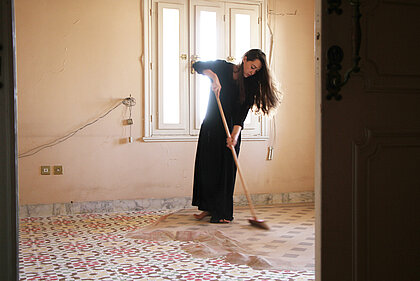 Eine Frau mit langem, dunklem Haar und schwarzem Kleid fegt über einen Boden, der von Sand bedeckt ist, in dem sich orientalische Muster abzeichnen.