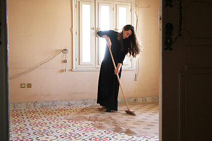 Eine Frau mit langem, dunklem Haar und schwarzem Kleid fegt über einen Boden, der von Sand bedeckt ist, in dem sich orientalische Muster abzeichnen.