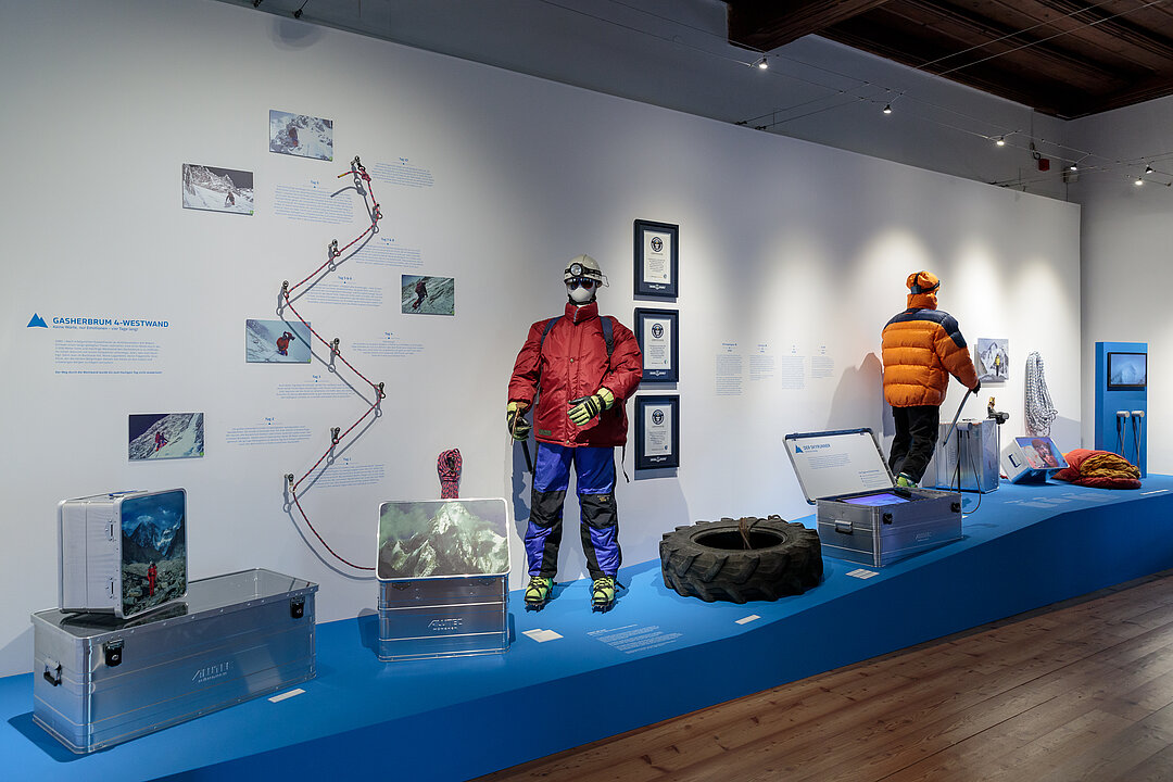 Ausstellungsansicht: Auf einem Podest stehen zwei Schaufenster-Puppen im Bergsteiger-Outfit. Daneben sind verschiedene Objekte und Ausstellungstexte zusehen.