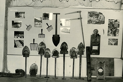 
Black and white photography of a wall. Various museum objects (mainly shovels) and photographs are attached to this.