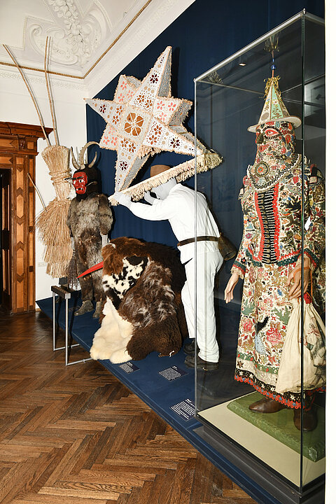 Photo of an exhibition room. On a long pedestal are various dressed up figures. One figure (Glöckler) wears a 1m high star on his head and shoulders. Another figure (Thomashutze) crouches on the floor and is covered with furs, only a red long beak can be seen.