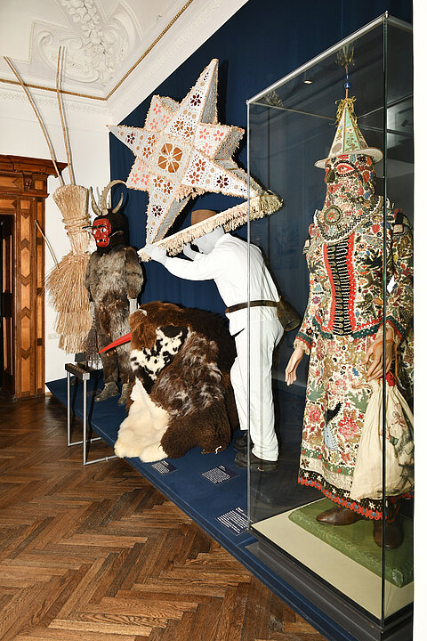 Foto von einem Ausstellungsraum. Auf einem langen Podest stehen verschiedene verkleidete Figuren. Eine Figur (Glöckler) trägt einen 1m hohen Stern auf seinem Kopf und den Schultern. Eine andere Figur (Thomashutze) kauert am Boden und ist mit Fellen bedeckt, nur ein Roter langer Schnabel ist zu sehen.