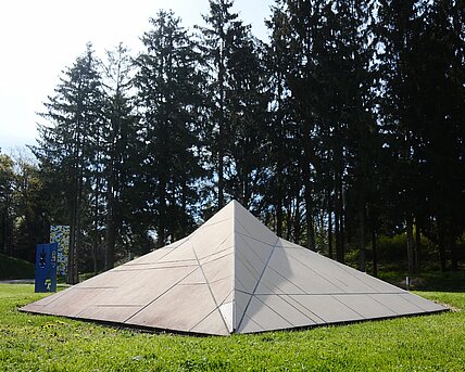 The sculpture in the shape of a stealth ship rests on the meadow. It is covered with beige-colored panels and cannot be detected by radar due to its shape.