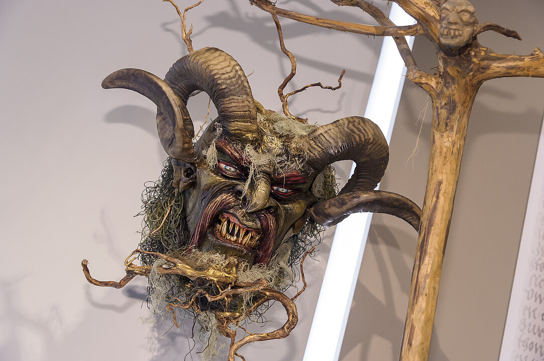 Eine Maske von einer Gestalt mit Hörnern und spitzen Zähnen hängt an einer Wand. An der Maske ist Moos und Äste befestigt.
