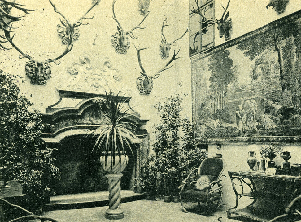 Photograph of a room with a fireplace. Antlers and a painting hang on the wall. There are plants everywhere