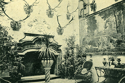 Fotografie von einem Zimmer mit Kamin. An der Wand hängen Geweihe und ein Gemälde. Überall stehen Pflanzen