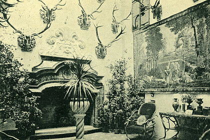 Photograph of a room with a fireplace. Antlers and a painting hang on the wall. There are plants everywhere