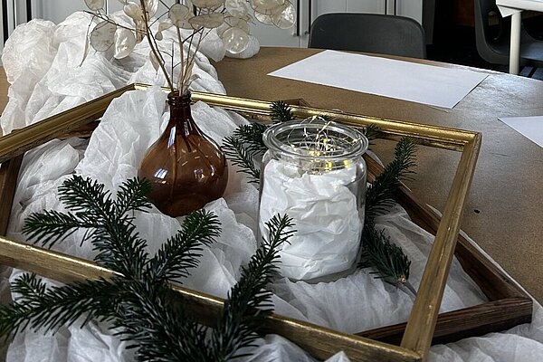 Tischdekoration mit brauner Vase und getrockneten transparenten Blättern, Glas mit Lichterkette, Tannenzweigen und goldenem Rahmen; im Hintergrund ein helles Atelier mit Tischen und Küchenzeile.