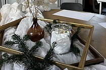 Tischdekoration mit brauner Vase und getrockneten transparenten Blättern, Glas mit Lichterkette, Tannenzweigen und goldenem Rahmen; im Hintergrund ein helles Atelier mit Tischen und Küchenzeile.