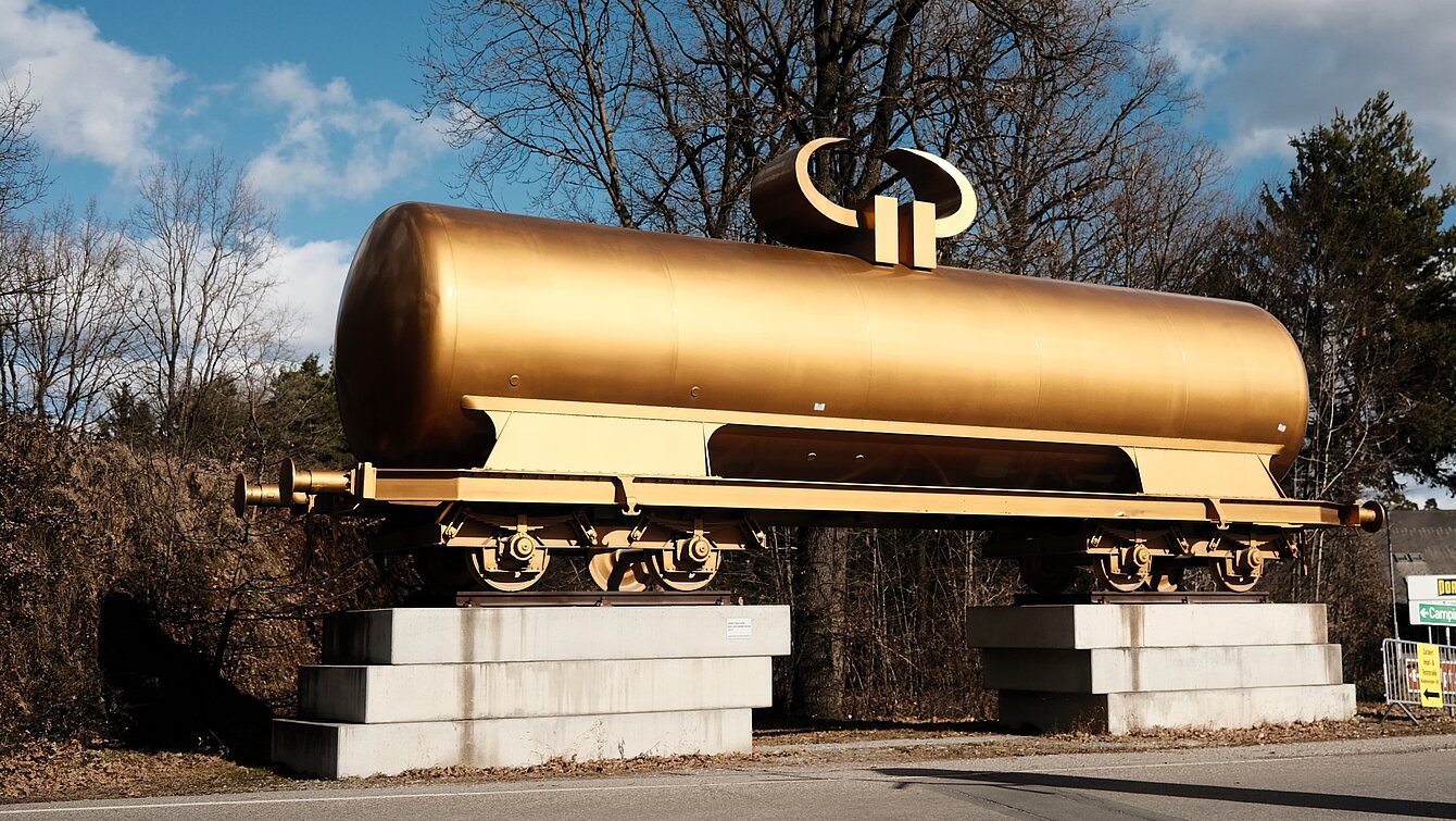 Ein goldener Kesselwagen für den Transport von Flüssigkeiten wie Mineralöl ist verziert mit einem liegenden Euro-Währungszeichen, welches die Figur zum gehörnten Götzenbild mutieren lässt. Sie befindet sich bereits vor dem Parkplatz außerhalb des Parkgeländes und lädt als erste Skulptur zum Besuch ein. 